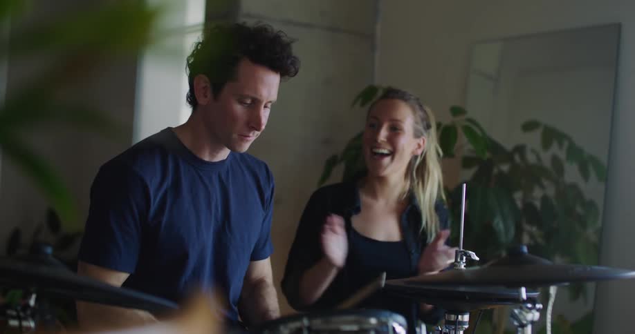 A man is holding drums sticks as he plays the drums and the woman sitting next to him is smiling as she cheers him on