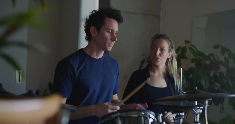 A man is holding drums sticks as he plays the drums as a blond woman sits smiling next to him