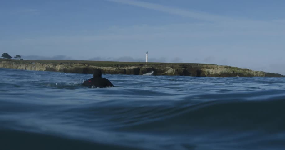 A man is lying on his surfboard as he paddles with his arms across the water towards shore