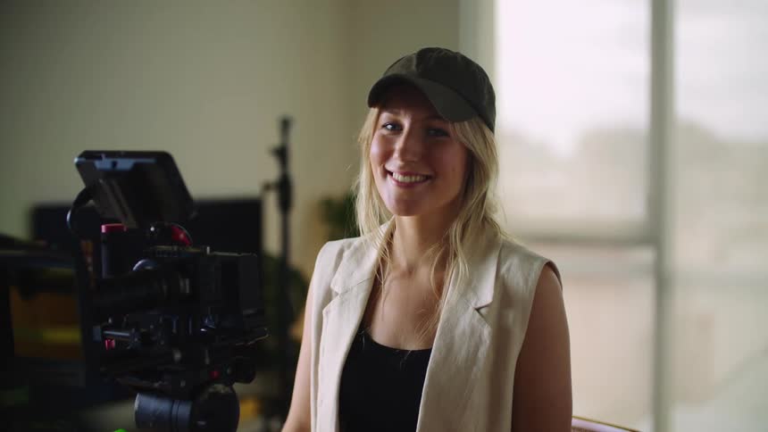 A blond woman wearing a black cap smiles as she stands behind a camera on a tripod