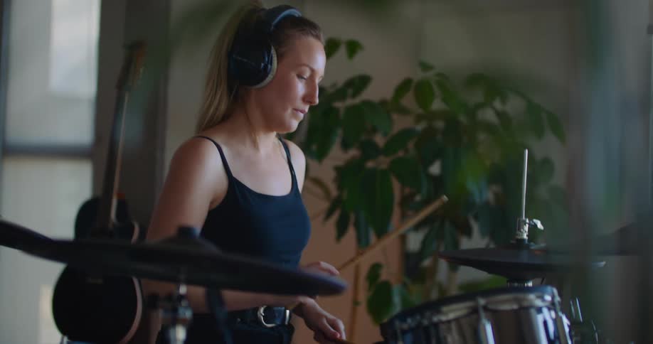 A blonde woman is practicing on a drum set indoors while wearing headphones