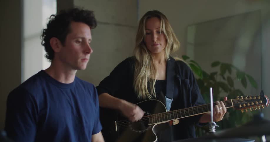 A man playing the drums and a woman playing a guitar next to him as they look at each other and smiling