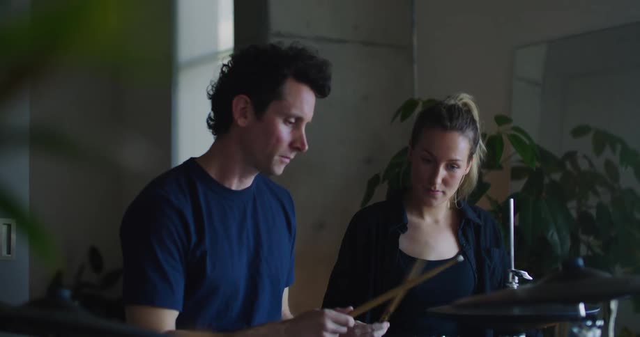 A man showing a woman how to hit the drum set with the drum sticks as she watches and nods