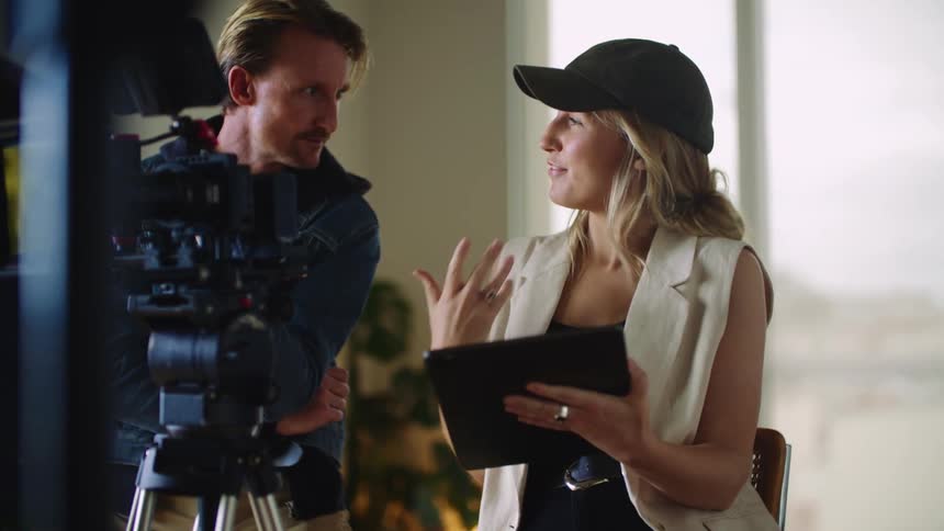 Blonde woman with cap black top and white vest talks to man wearing blue coat behind movie camera