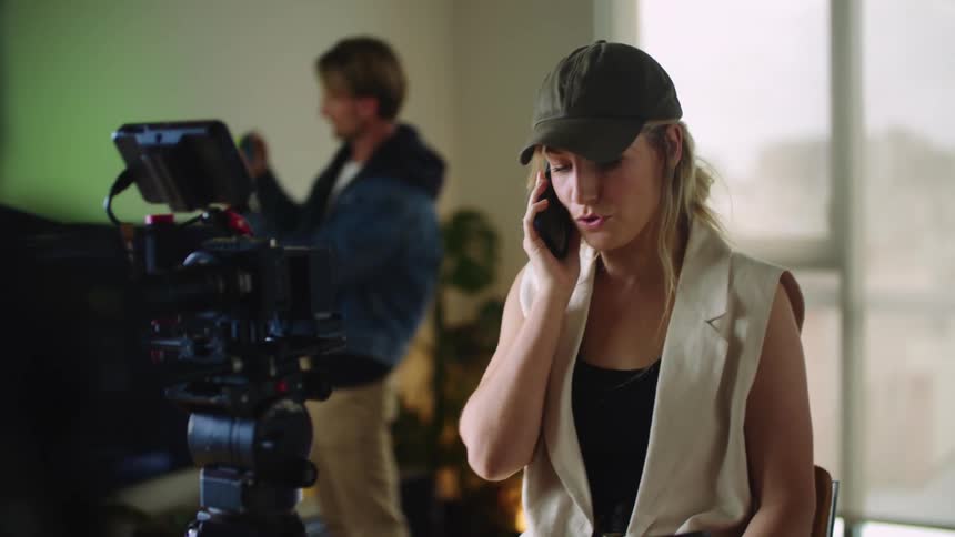 Woman with cap black top and white vest talks on mobile phone next to movie camera