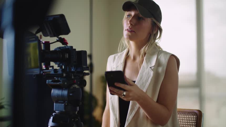 A woman is standing behind a camera on a tripod as she holds her phone and dials with her thumb