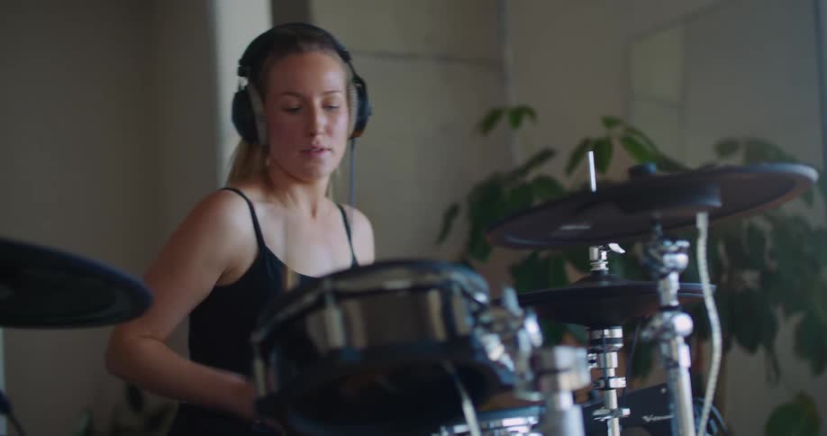 Blonde woman with ponytail headphones and black shirt plays drums in room