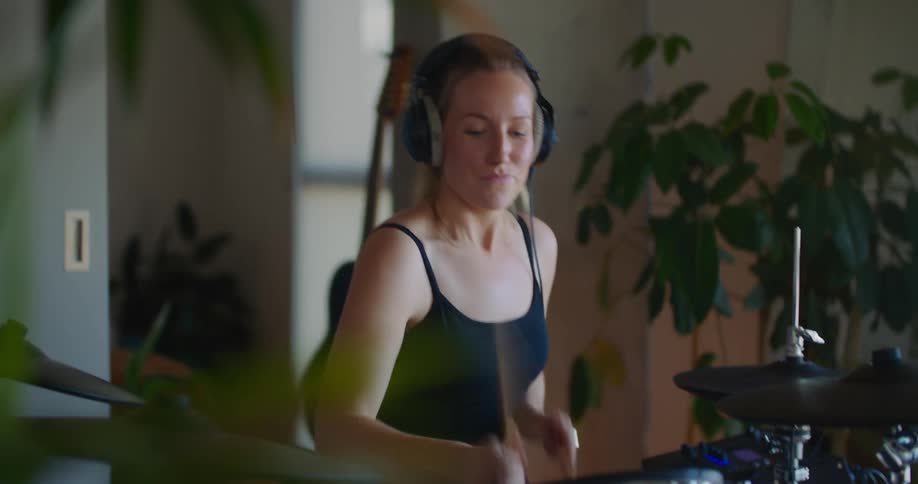 Blonde woman with ponytail black top and headphones play drums in room