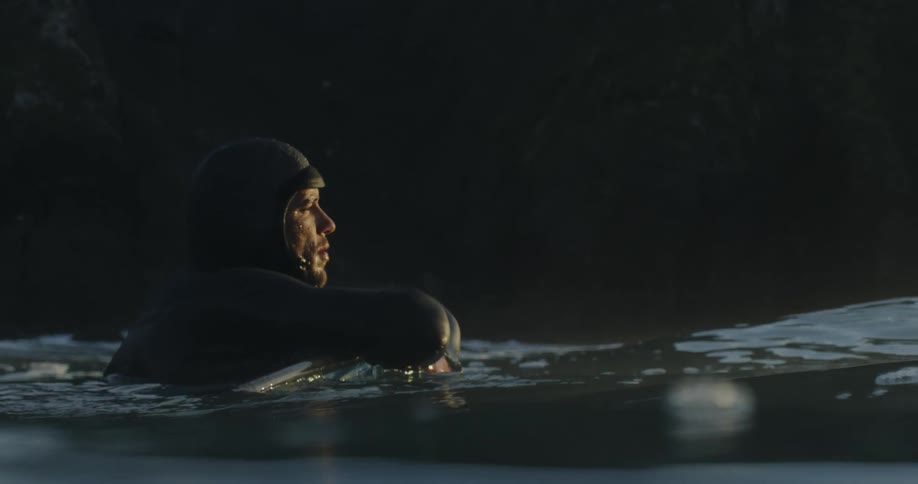 A man wearing a wet suit floating in the water hanging onto a board while looking out over the ocean