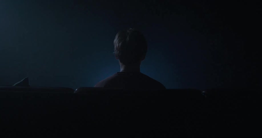A man is sitting on a couch looking at a flashing light or TV in the darkness
