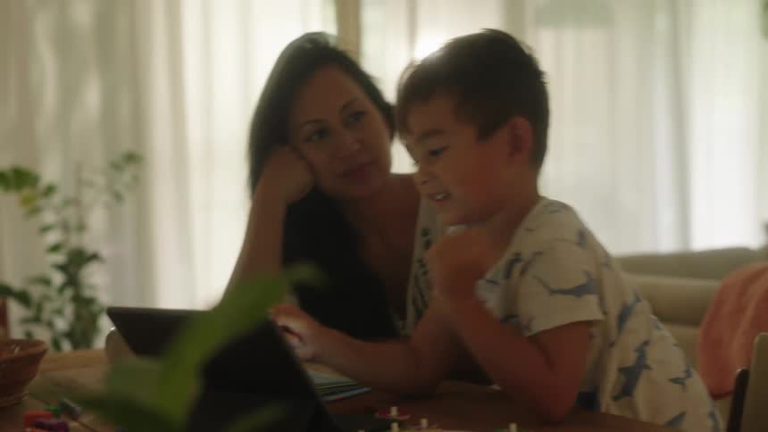Mother and son sitting together at dining table laughing and smiling reading probably home schooling