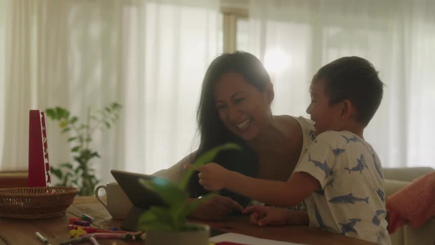 A boy and his mom laugh while watching something on a tablet at their dining room table