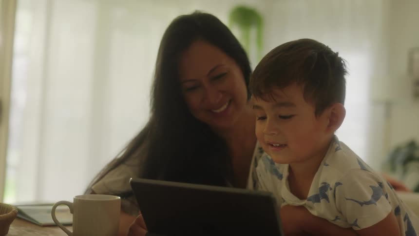 A mother and son look at a tablet together as the mother smiles while the boy reads on the tablet