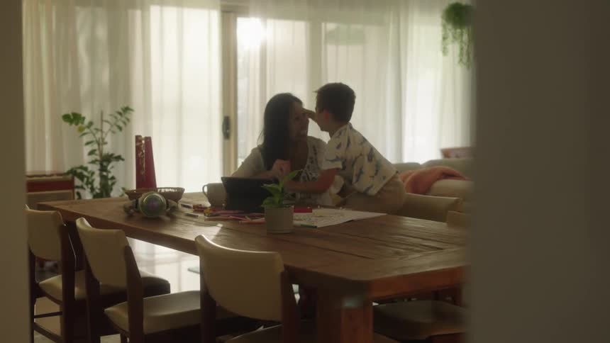 A boy stands up to hug his mom while sitting in front of a laptop