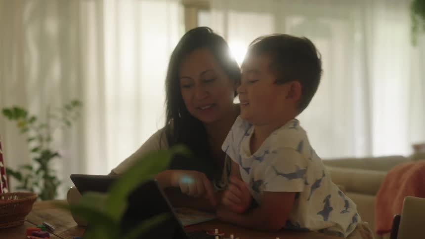 Mother and toddler son sitting at dining table working on laptop together probably home schooling laughing and smiling
