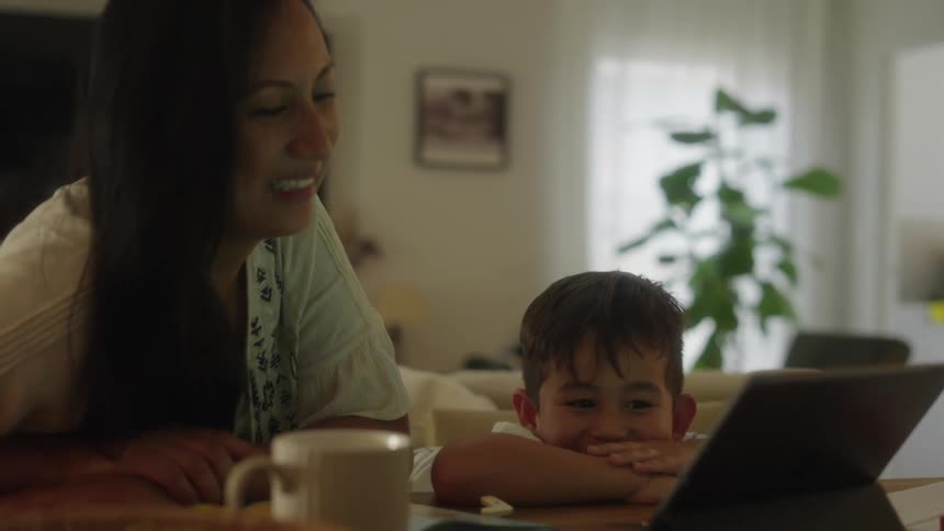 Mother and son sitting together at dining table with tablet laughing and smiling probably home schooling