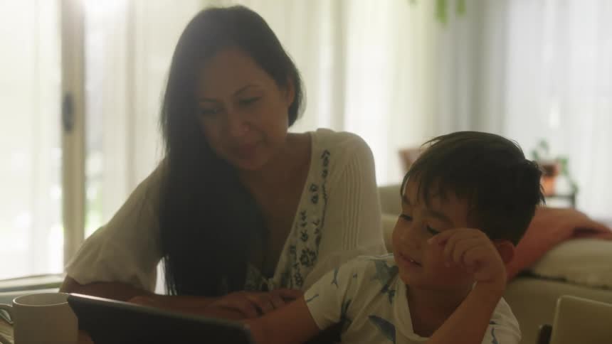 A mother and her son sitting at a table looking at a tablet smiling and laughing