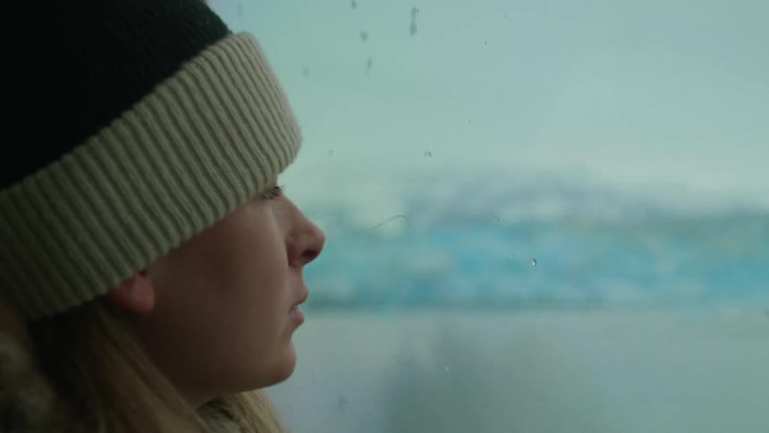 Blonde woman with wooly hat looks out the window of a boat traveling on water next to snowy mountain