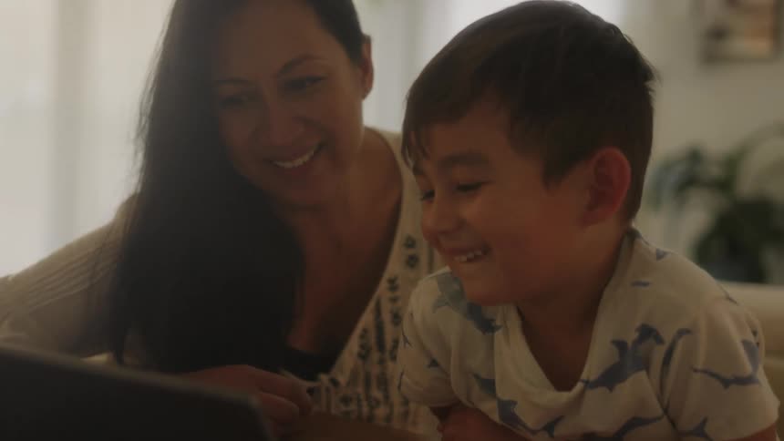 A laughing boy watches the screen of a laptop alongside his mother in the living room of a house during the day