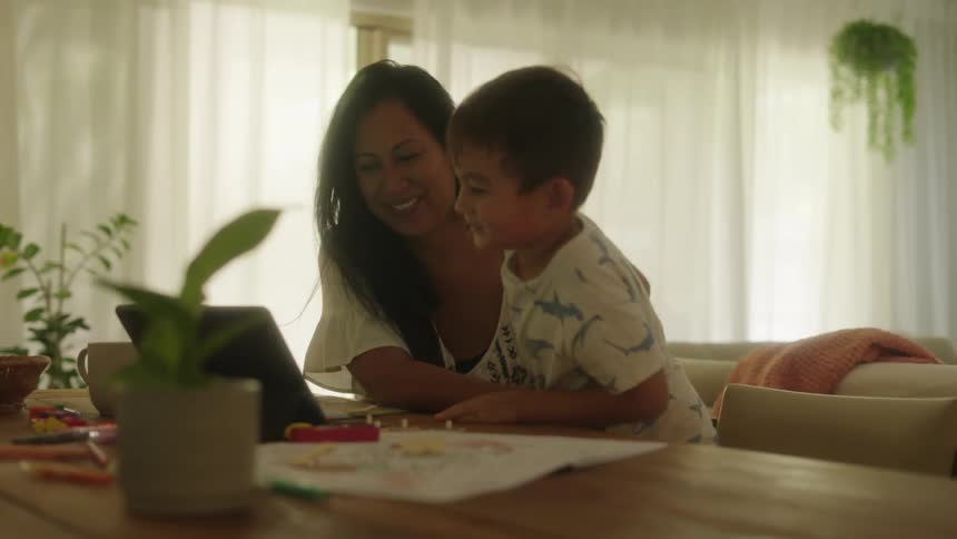 Mother and son at home sitting at dining table with laptop drawing materials and toys probably home school