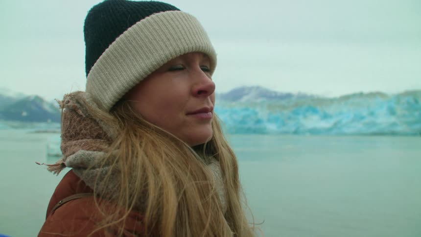 Blonde woman with wooly hat and brown coat watches the lake and glacier