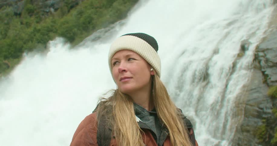 A woman standing near a waterfall in the wilderness turns to gaze around at her surroundings