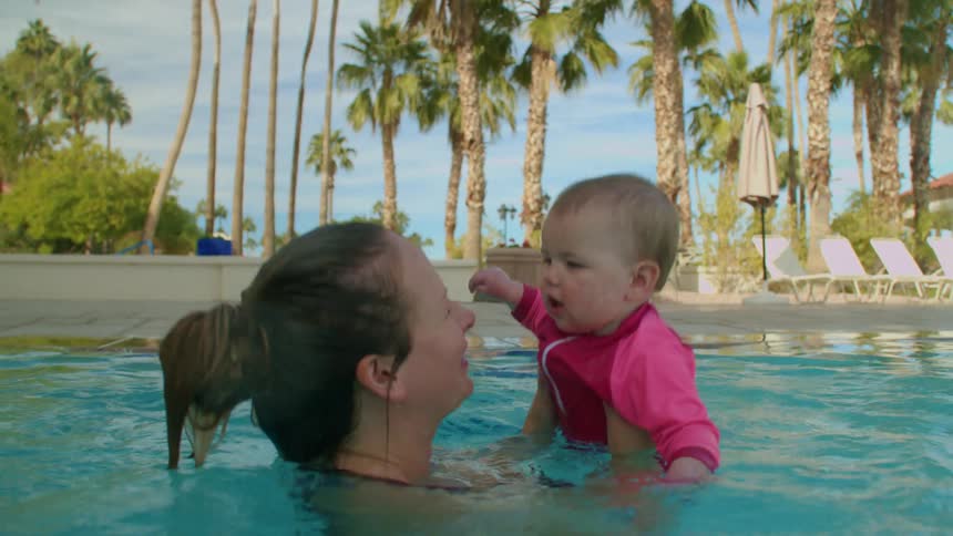 A mother throws her baby into the air in a swimming pool as the baby blinks at the mother