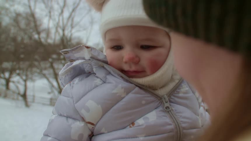 Mother with green wooly hat brown jacket holds baby with white hat and purple coat in her arms in the snowy public park