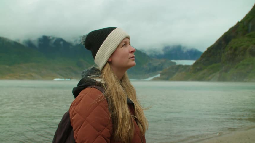 Blonde woman stands on beach with her back to the water while she looks up and low lying clouds cover mountains