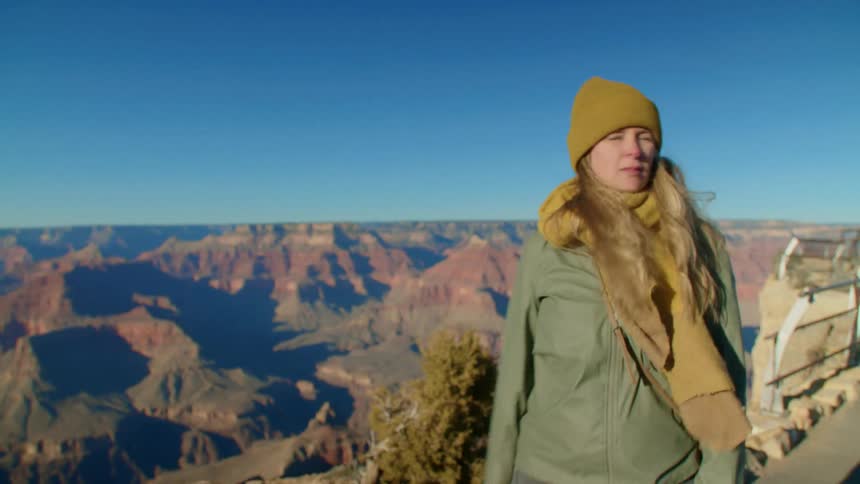 A woman wearing a yellow scarf and hat stands at the edge of a cliff turning around and looking around her