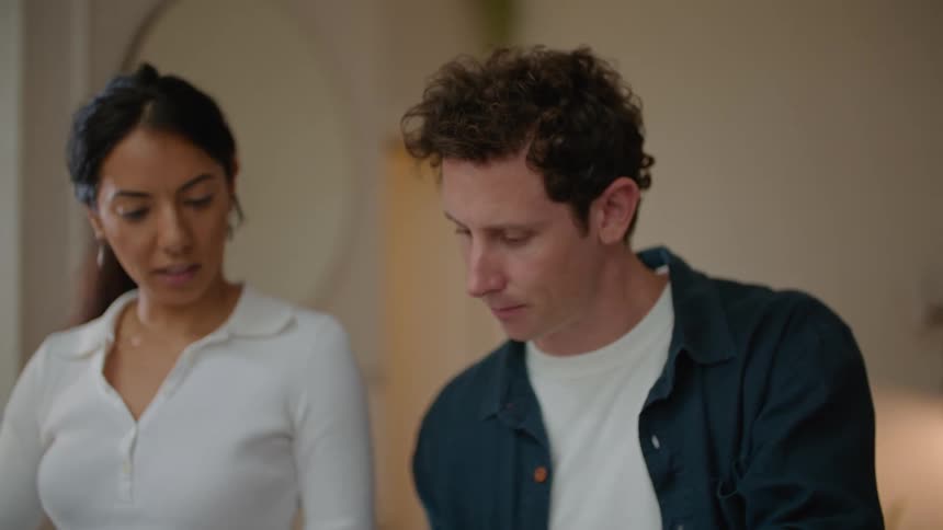A woman is standing at a table scrolling through pictures on her laptop computer as she talks to a man sitting next to her