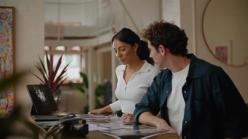 A man is sitting at a desk with pictures in front of him as he looks at a laptop computer with the woman standing next to him