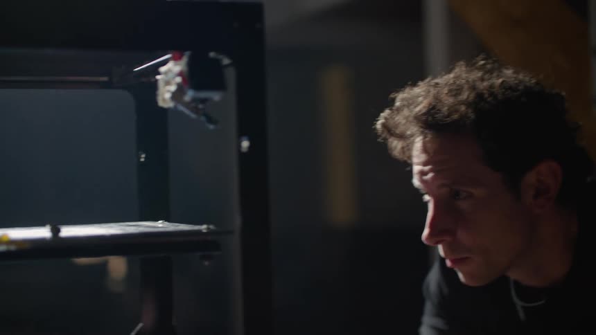 Man in warehouse standing in front of a 3d printer observing and finetuning adjusting dials