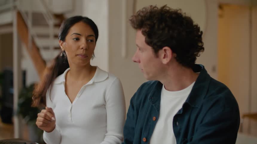A man is sitting at a table holding a pen and writing as the woman sitting next to him is working on a laptop computer as they talk and she laughs