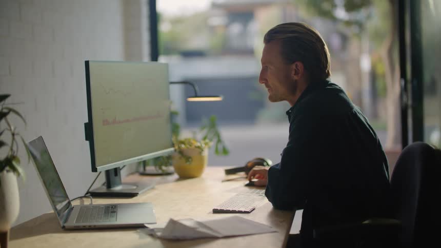 Man sits in office with view of road and references both large monitor and laptop