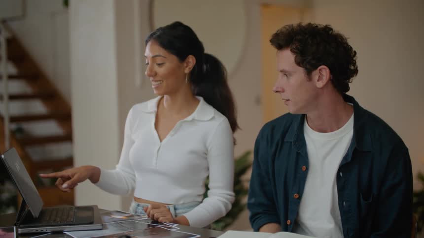 A man is sitting at a table holding a pen as he looks at a laptop computer with the woman standing next to him