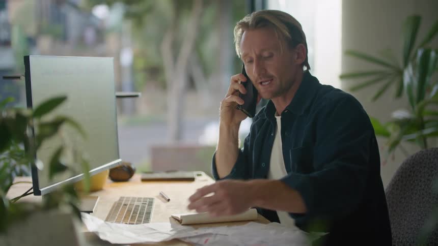 A man sitting at his desk talking on his cell phone looking at a paper smiling and nodding his head