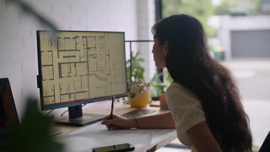 Woman makes notes on paper while consulting plan on computer monitor