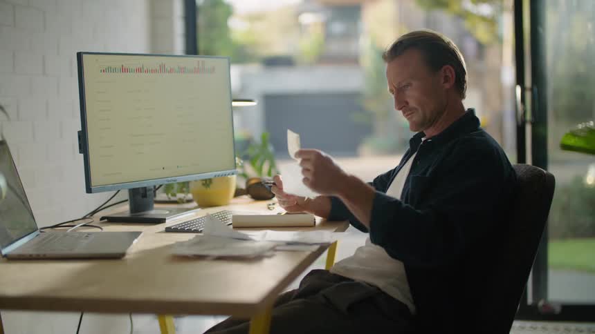 A man sitting at his desk in front of a computer screen and laptop while looking through papers and typing on the computer