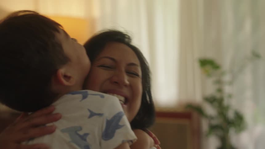 A mother smiles and hugs her son next to her as he stands upt and falls into her arms