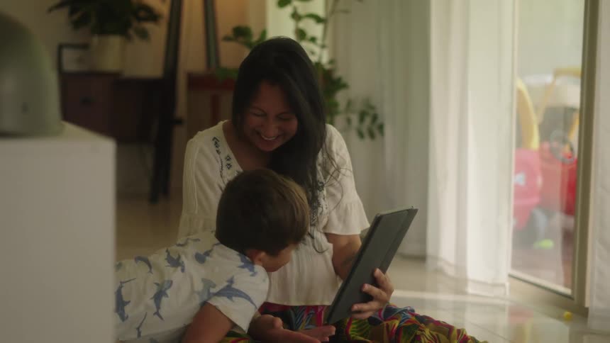 Mother with white shirt and colorful pants uses tablet with son sitting next to her in the living room