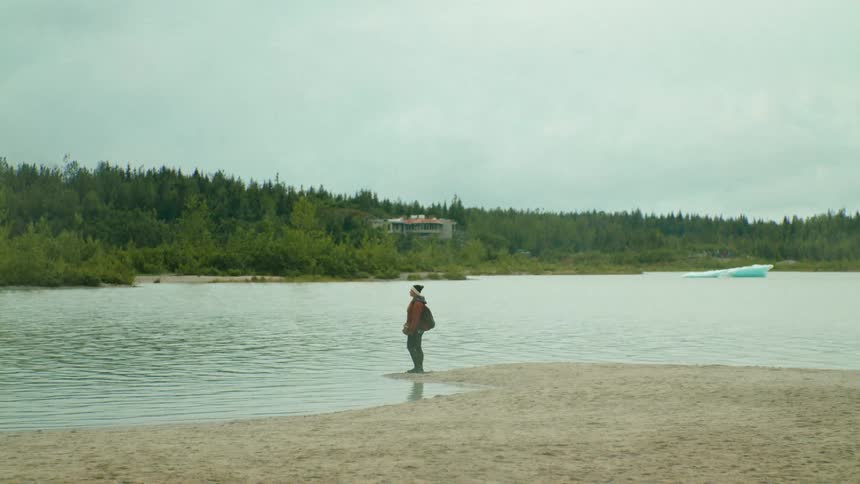 Woman with wooly hat red coat black backpack and pants stands on the bank of a lake watching the view on a cloudy day