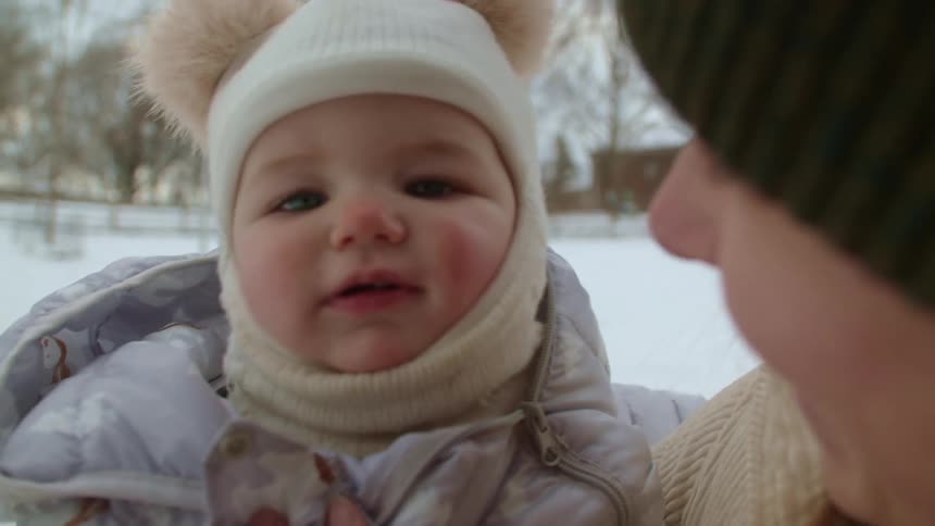 Mother with green wooly hat and beige coat holds baby with white hat and purple coat in the snowy field