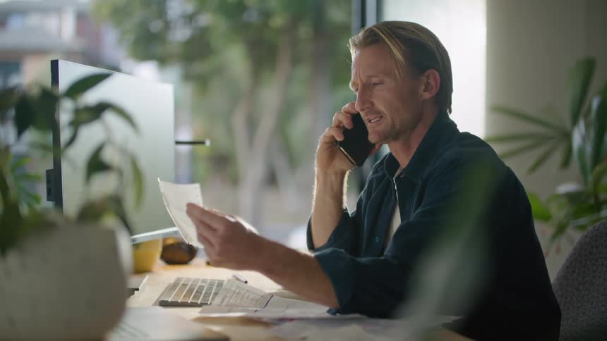 Man discusses contents of paper on phone while sitting at desk with a computer