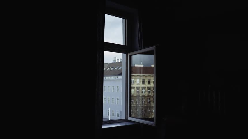 An open window in a dark room reveals old weathered buildings in the city