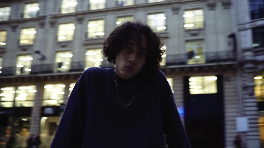 A teenage boy sitting on a city railing on a skatebaord looking aggressive
