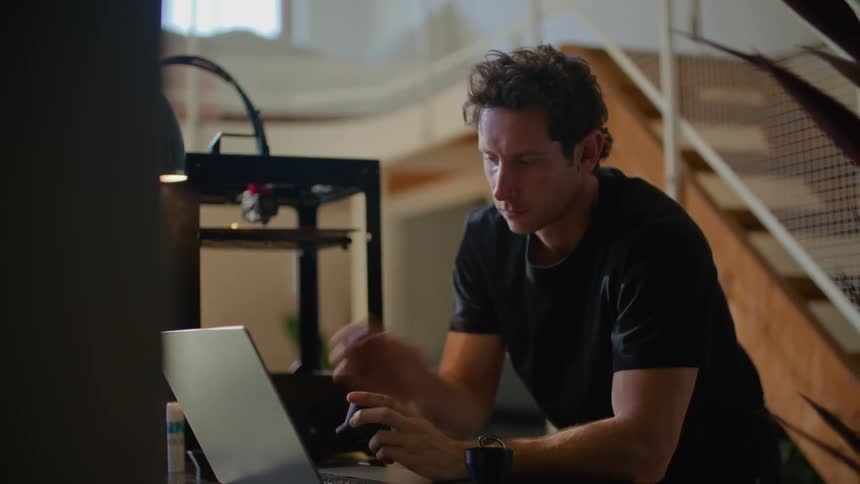 Man types on laptop near 3d printer and stops to examine object