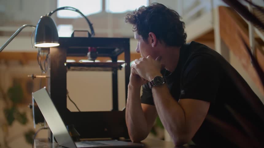 A man is sitting at a desk with a laptop computer with his hands on his face as he thinks and a machine moves near him