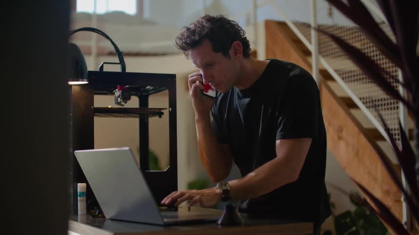 Businessman in black t shirt and wristwatch talks on smartphone and uses laptop at the office