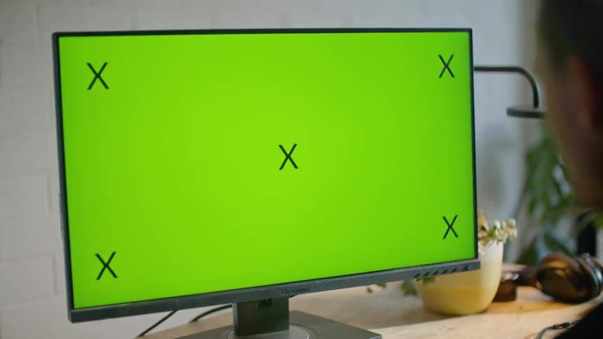 A man is sitting at a desk looking at a computer with a green screen